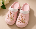 Pink clogs with decorative charms on a beige surface with flowers in the background