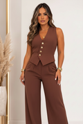 A woman models a sleeveless brown jumpsuit with a structured corset-style bodice, decorative gold buttons down the front, and matching wide-leg pants. She is standing in a brightly lit indoor setting with a neutral wall and a decorative vase of pampas grass in the background.