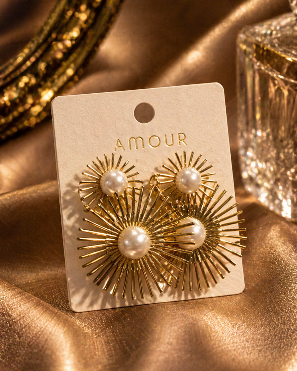 Gold earrings with pearl centers on a 'AMOUR' branded card against a gold fabric background