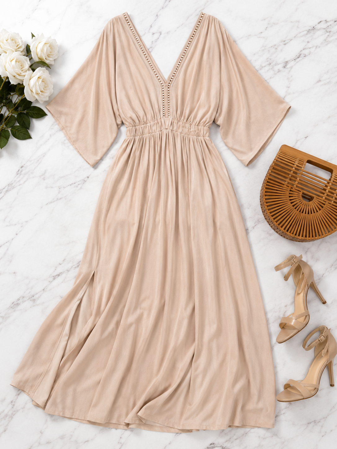 A beige maxi dress with elbow-length sleeves, a V-neckline, and delicate crochet trim along the neckline and waist, styled in a flat lay on a white marble background with white roses, a woven clutch, and nude high heels.