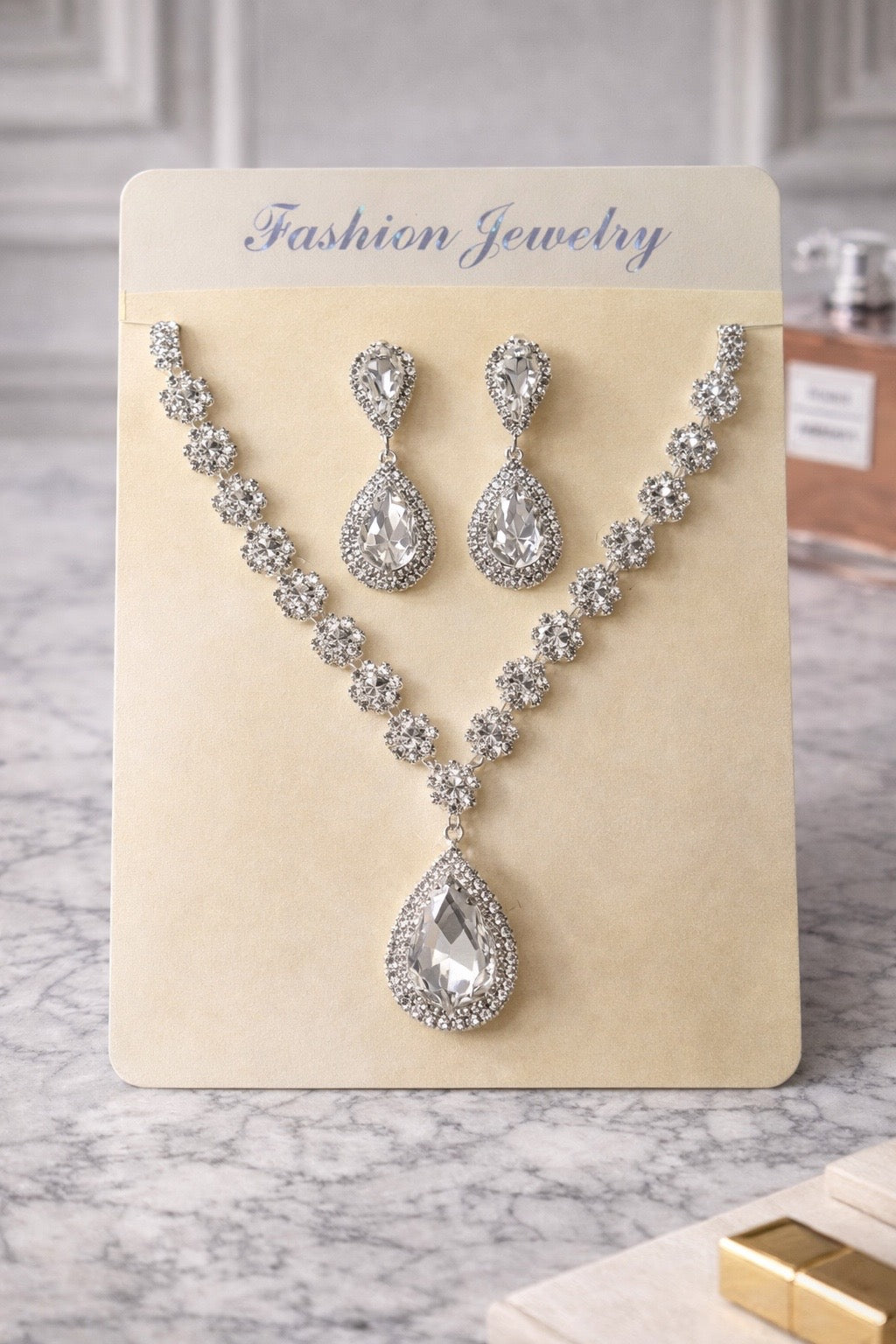 Silver jewelry set with necklace and earrings on a beige card labeled 'Fashion Jewelry' against a marble background.