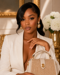 A woman in a white blazer holds a beige rhinestone-embellished handbag with gold hardware in an elegant indoor setting with gold-framed wall art and white flowers.