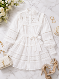 A white long-sleeve lace blouse with a high collar, ruffled neckline, and matching waist tie, styled in a flat lay on a marble surface with flowers, shoes, jewelry, and a handbag.