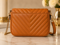 A tan quilted leather shoulder bag with a chevron pattern, featuring a gold chain strap and a matching leather strap, displayed on a marble surface with a blurred background.