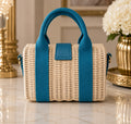 A small woven straw tote bag with bright teal blue handles, side straps, and a front flap detail, displayed on a polished marble surface with decorative gold and white floral elements in the background.