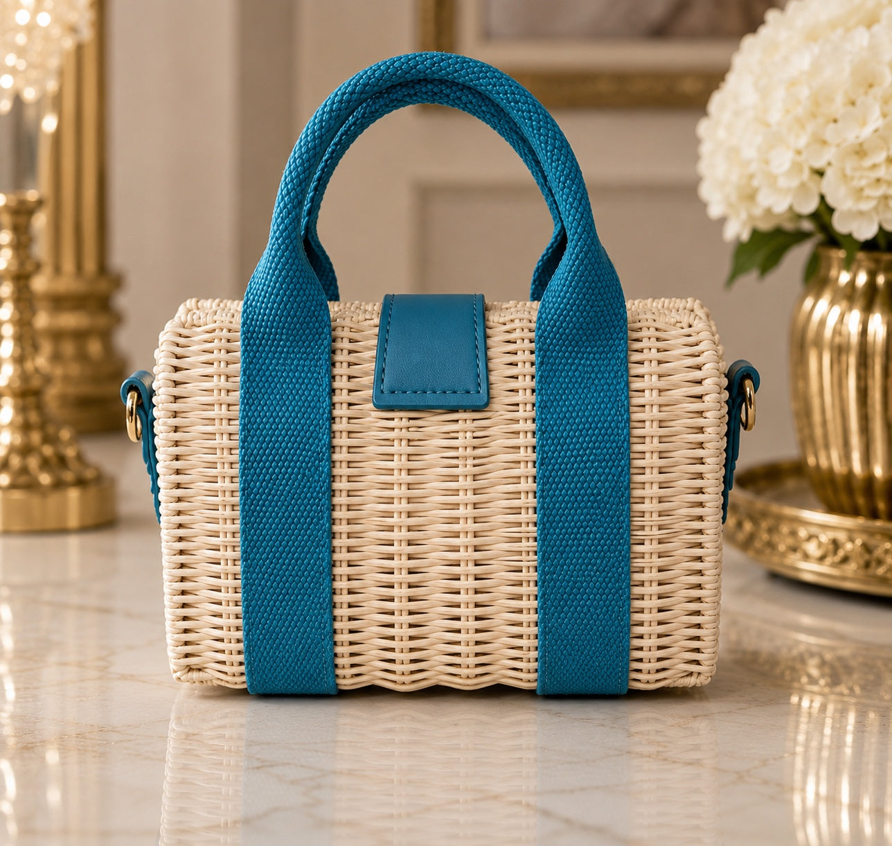 A small woven straw tote bag with bright teal blue handles, side straps, and a front flap detail, displayed on a polished marble surface with decorative gold and white floral elements in the background.