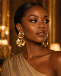 A woman wearing large, chunky gold double-drop statement earrings with a faceted, textured finish, set against a warm, elegant background with soft golden lighting.