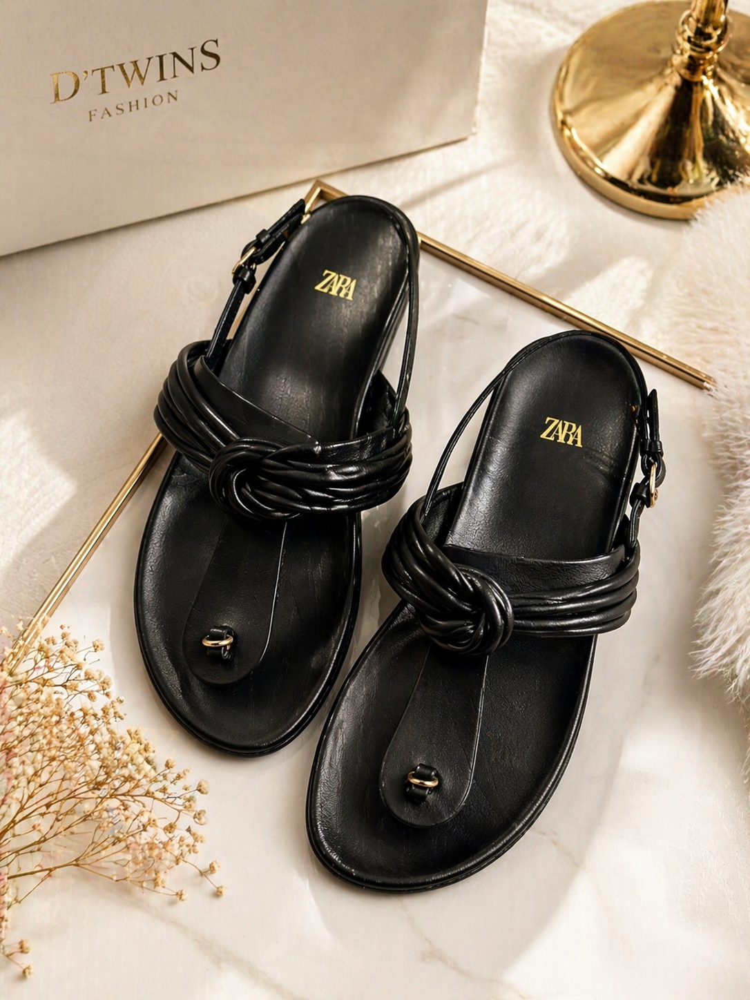 Black sandals with braided straps on a light surface with 'D'Twins' branding.
