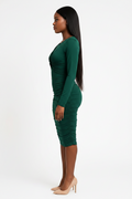A woman in profile wearing a fitted long-sleeve ruched midi dress in emerald green, paired with nude high heels, against a plain white studio background.