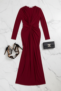 A long-sleeve floor-length dress in deep burgundy red, styled in a flat lay with black strappy high-heeled sandals and a black clutch bag on a white marble background.