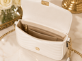A white quilted shoulder bag with gold-tone hardware, shown open to reveal a beige interior with card slots and a zip pocket. The bag is styled on a light marble surface with decorative gold and white flowers in the background.