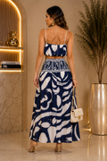 A woman stands with her back to the camera wearing a navy and white abstract-patterned two-piece set consisting of a crop top and a maxi skirt. She is holding a small white handbag in a luxury indoor setting with warm lighting.