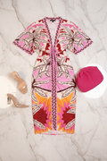 A short-sleeve wrap dress with a vibrant pink, orange, and brown palm tree pattern, styled in a flat lay with beige high-heeled sandals and a pink knit hat on a white marble background.