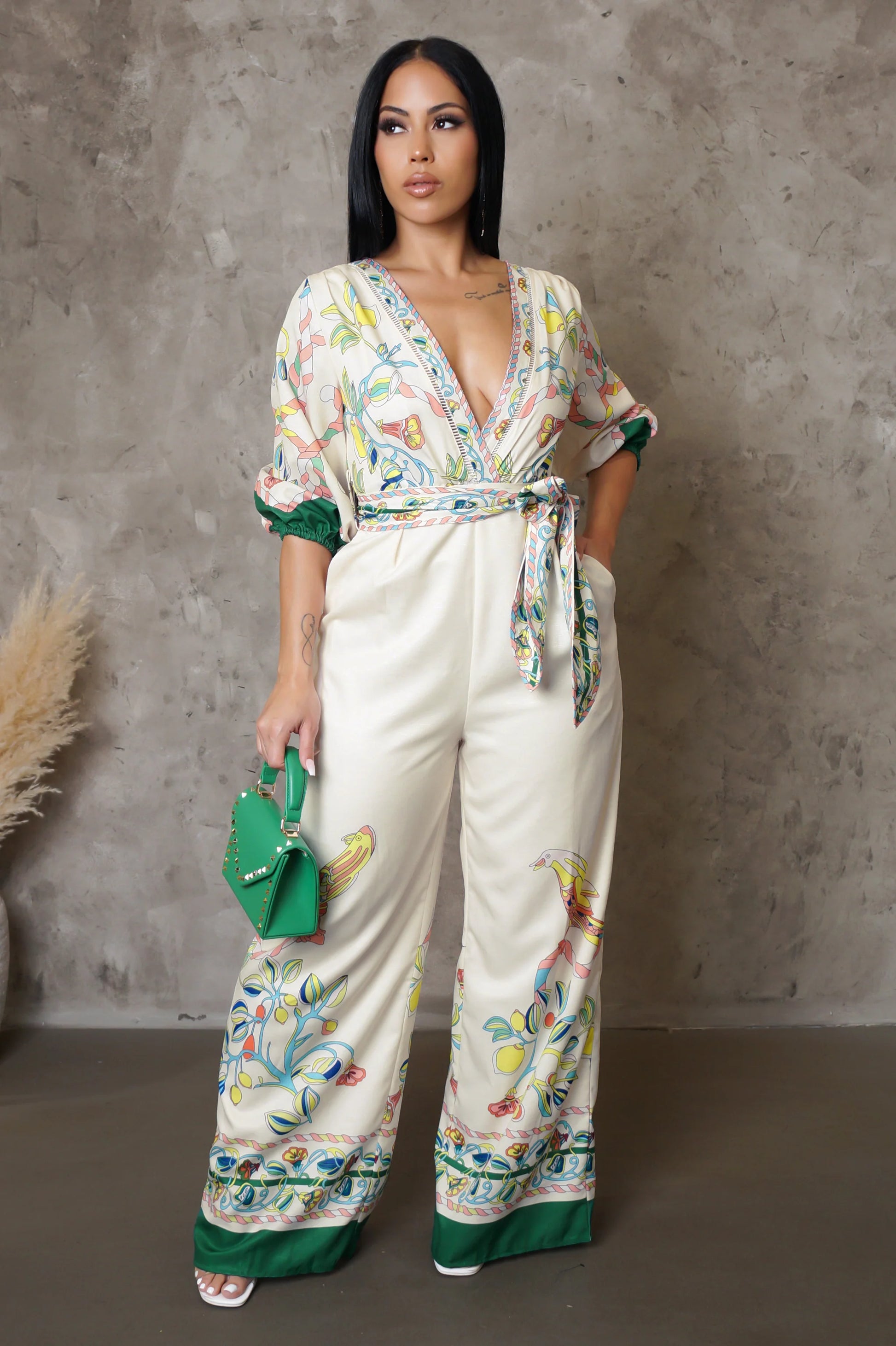 Woman wearing a floral jumpsuit with a green belt and holding a green handbag against a gray wall.