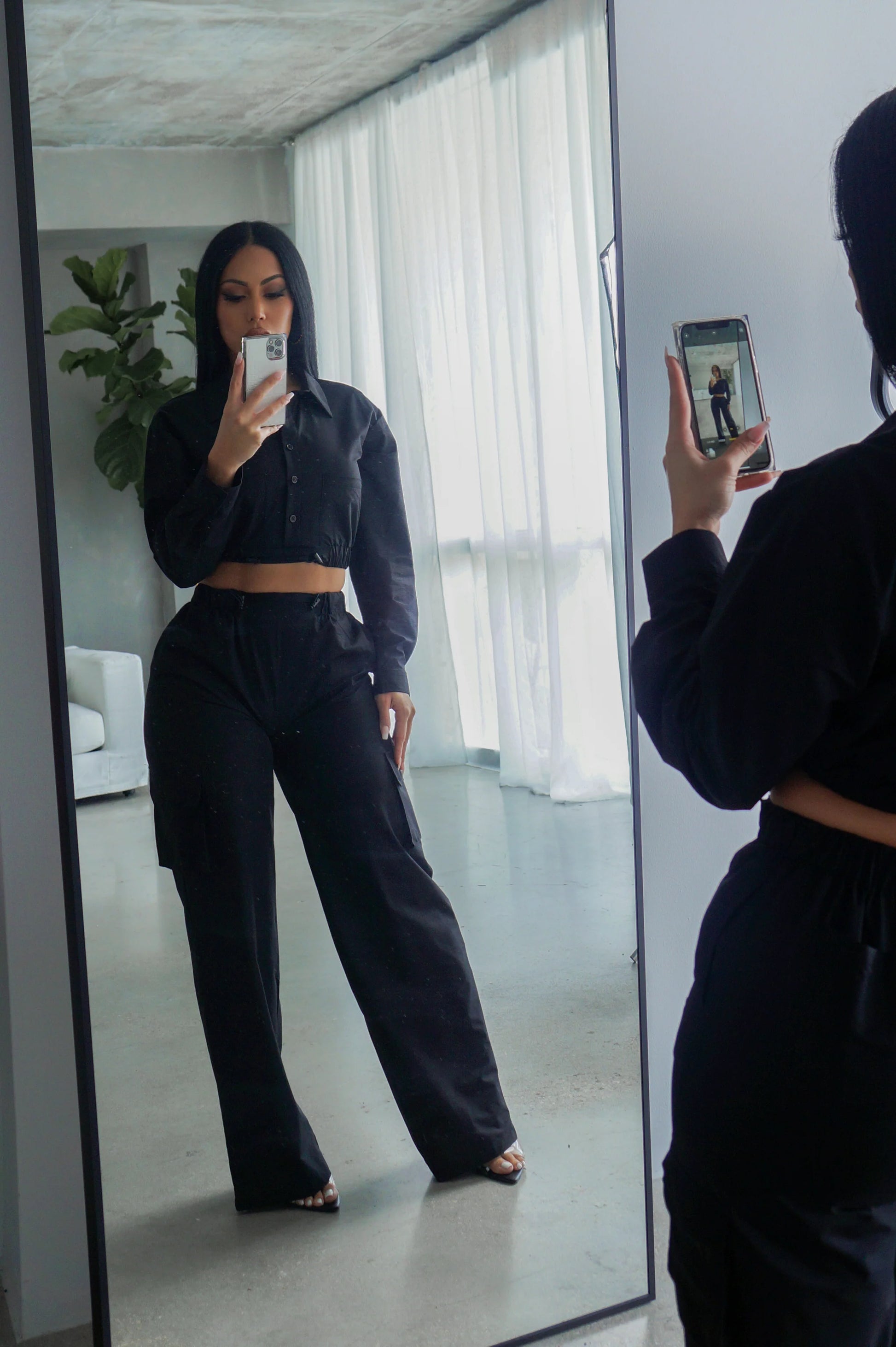 Person taking a mirror selfie wearing a black outfit in a modern room.