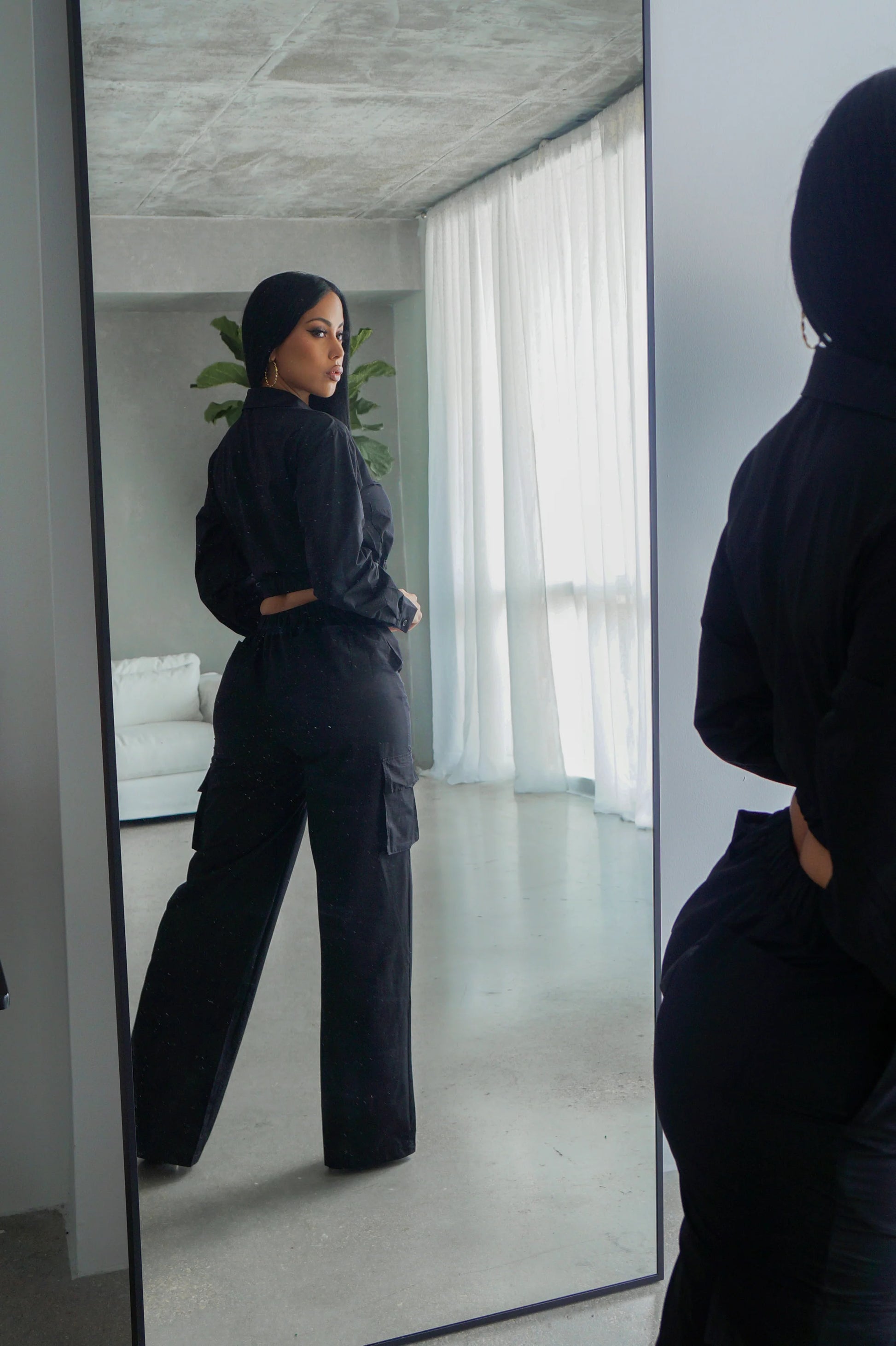 Person wearing a black outfit standing in front of a mirror in a modern interior setting.