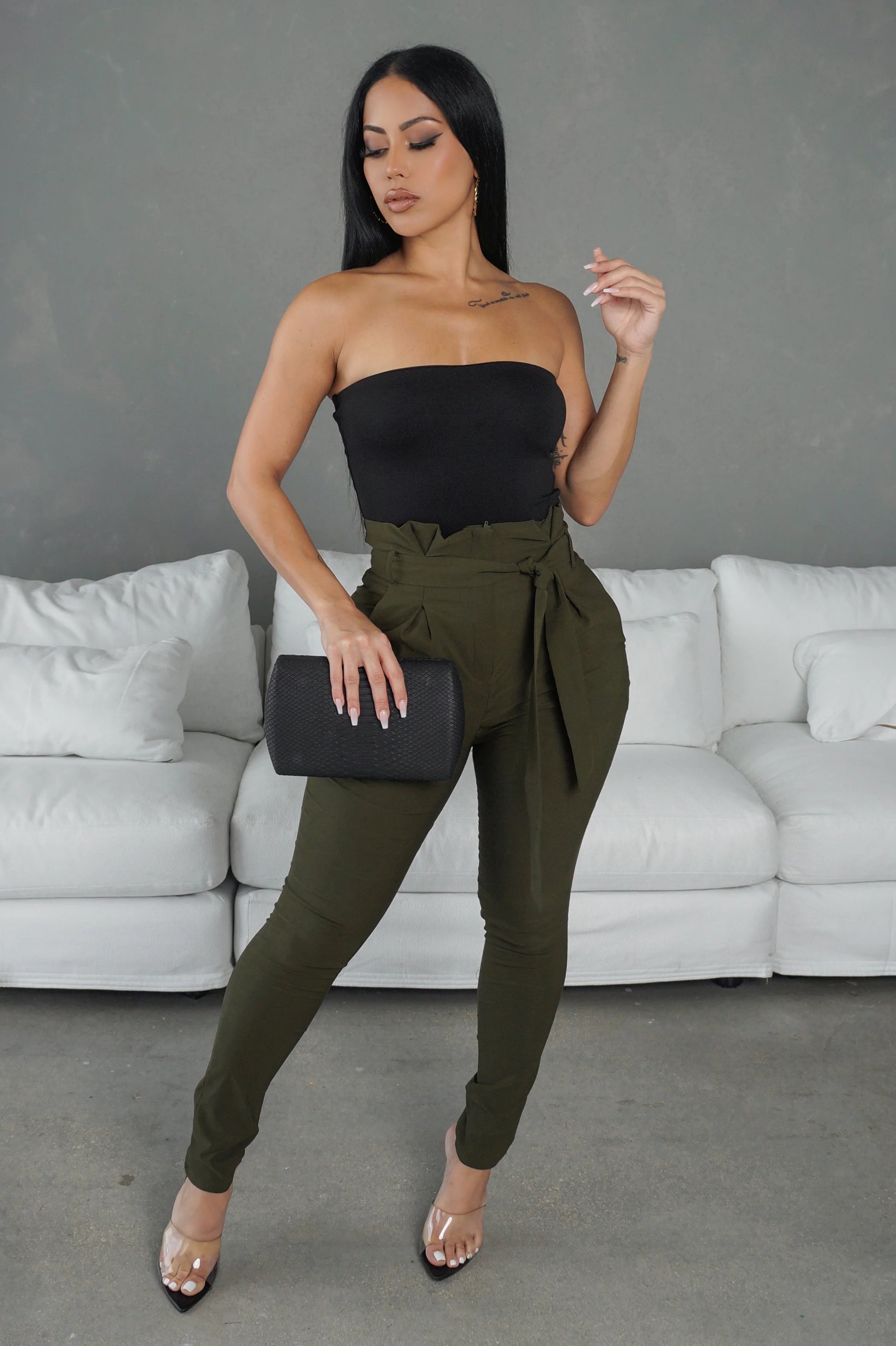 Woman in black top and green pants posing in front of a gray wall with a white couch.