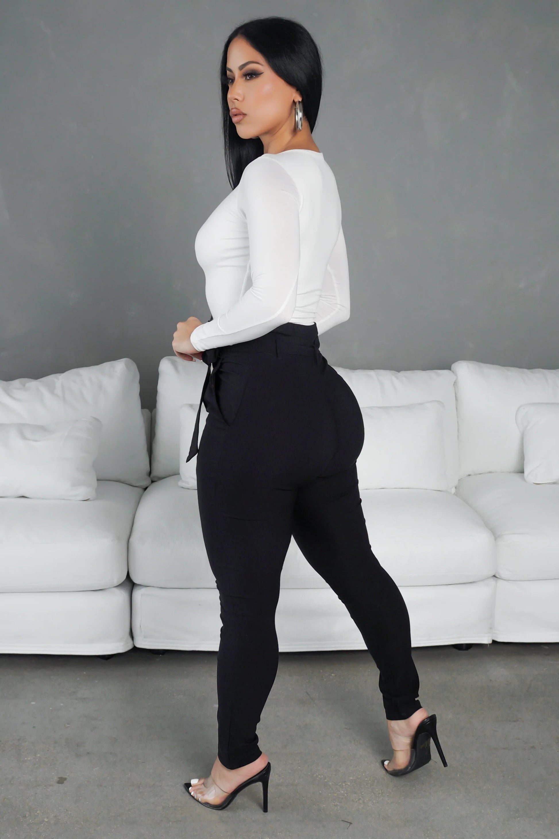 Woman in a white top and black pants standing in front of a white sofa with a gray wall background.