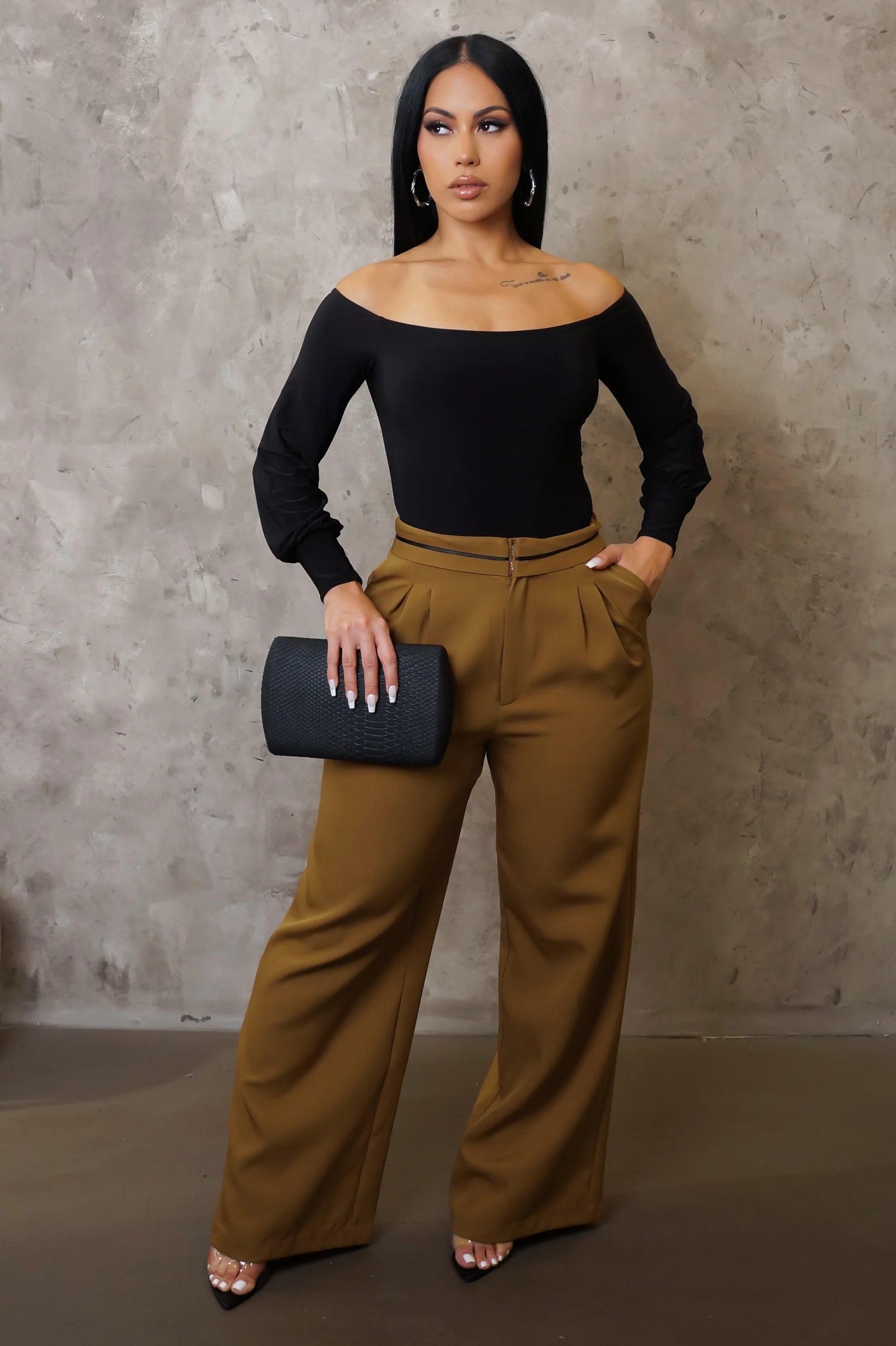 Woman wearing a black top and brown pants holding a clutch against a neutral background