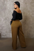 Woman wearing a black off-shoulder top and mustard yellow pants against a textured wall.