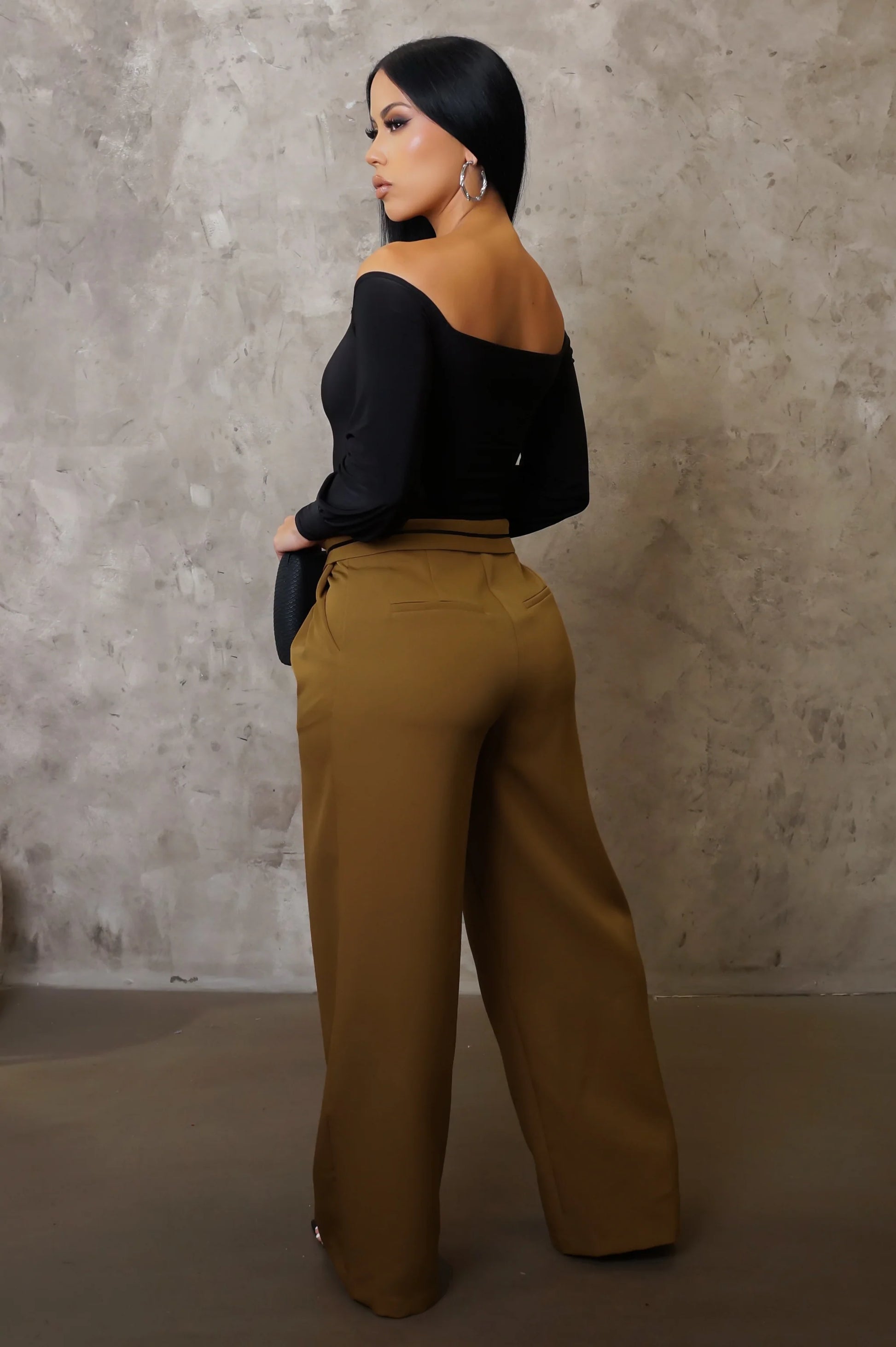 Woman wearing a black off-shoulder top and mustard yellow pants against a textured wall.
