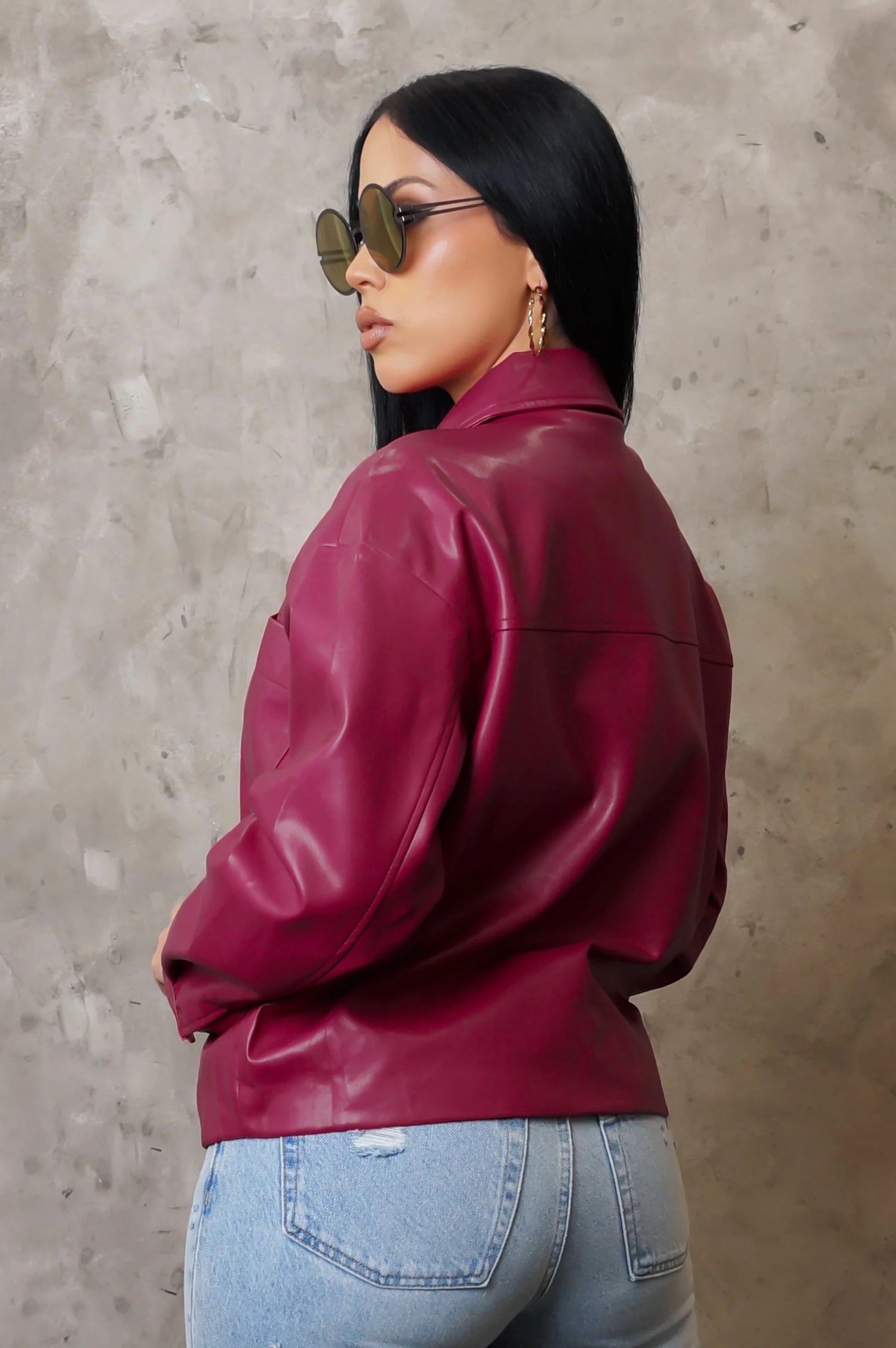 Woman wearing a burgundy leather jacket and light blue jeans against a beige wall.