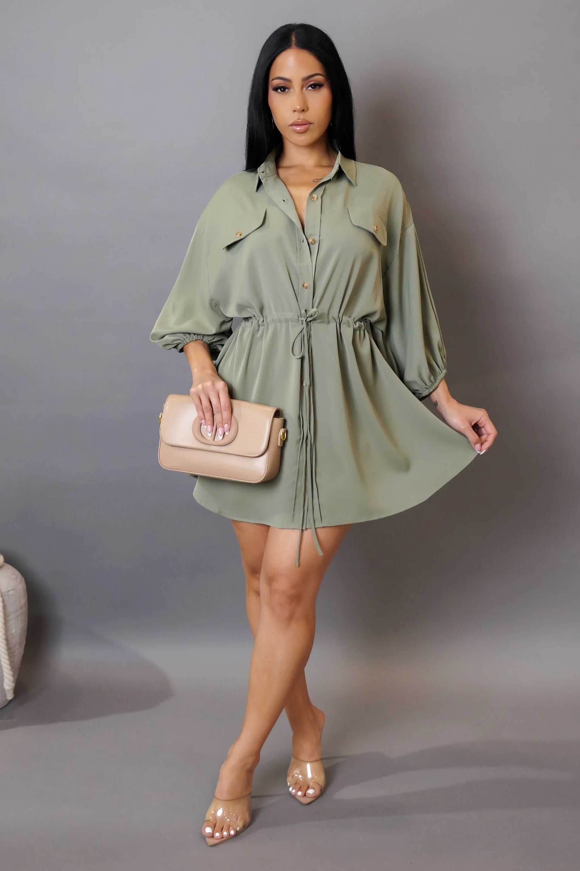 Woman wearing a green dress holding a beige handbag against a gray background