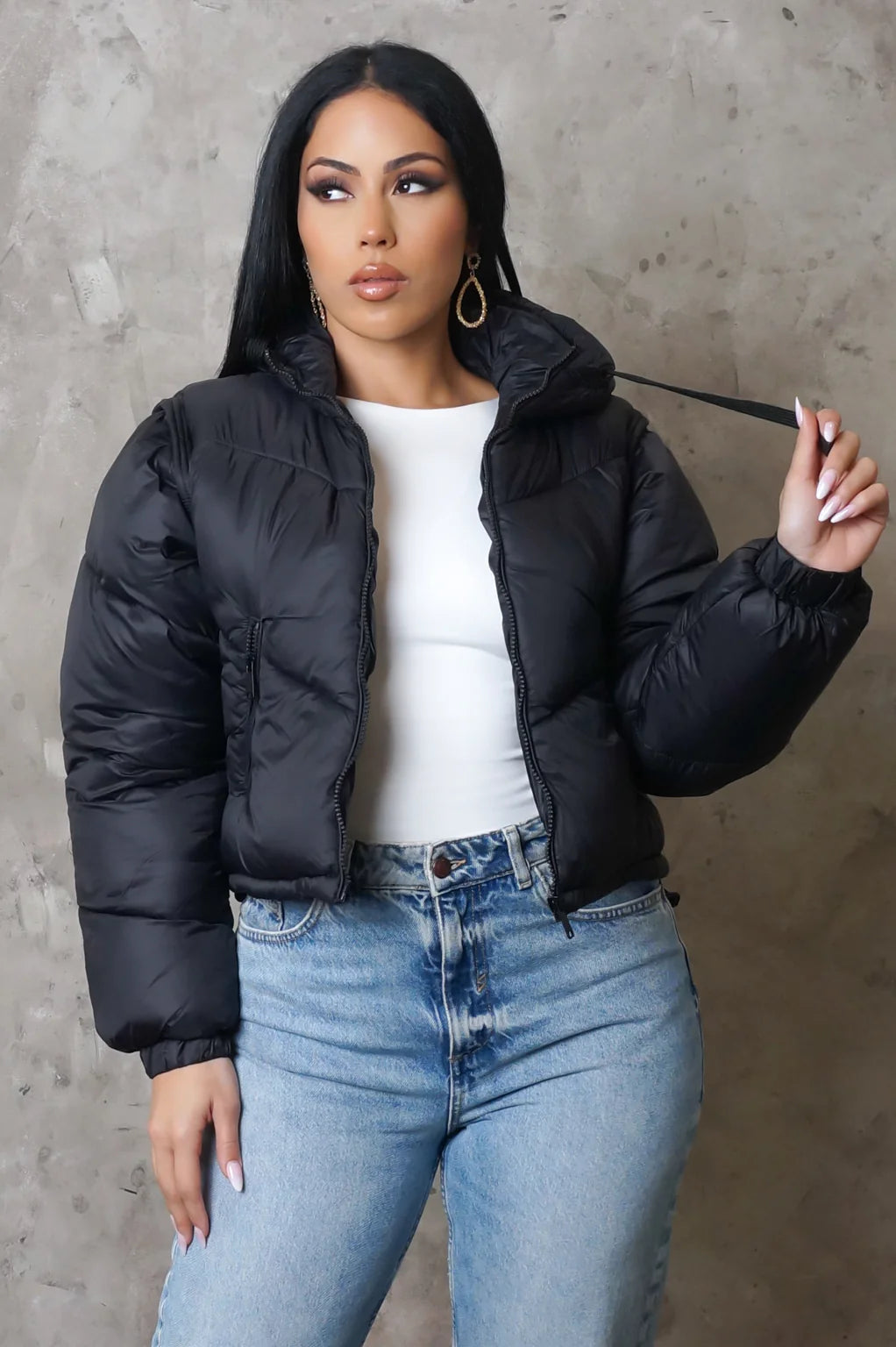 Woman wearing a black puffer jacket and blue jeans against a gray concrete wall.