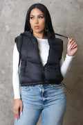 Woman wearing a black puffer vest over a white top and blue jeans against a gray concrete wall.