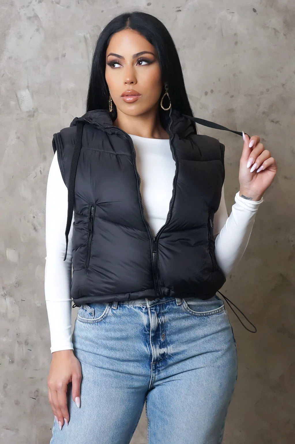 Woman wearing a black puffer vest over a white top and blue jeans against a gray concrete wall.