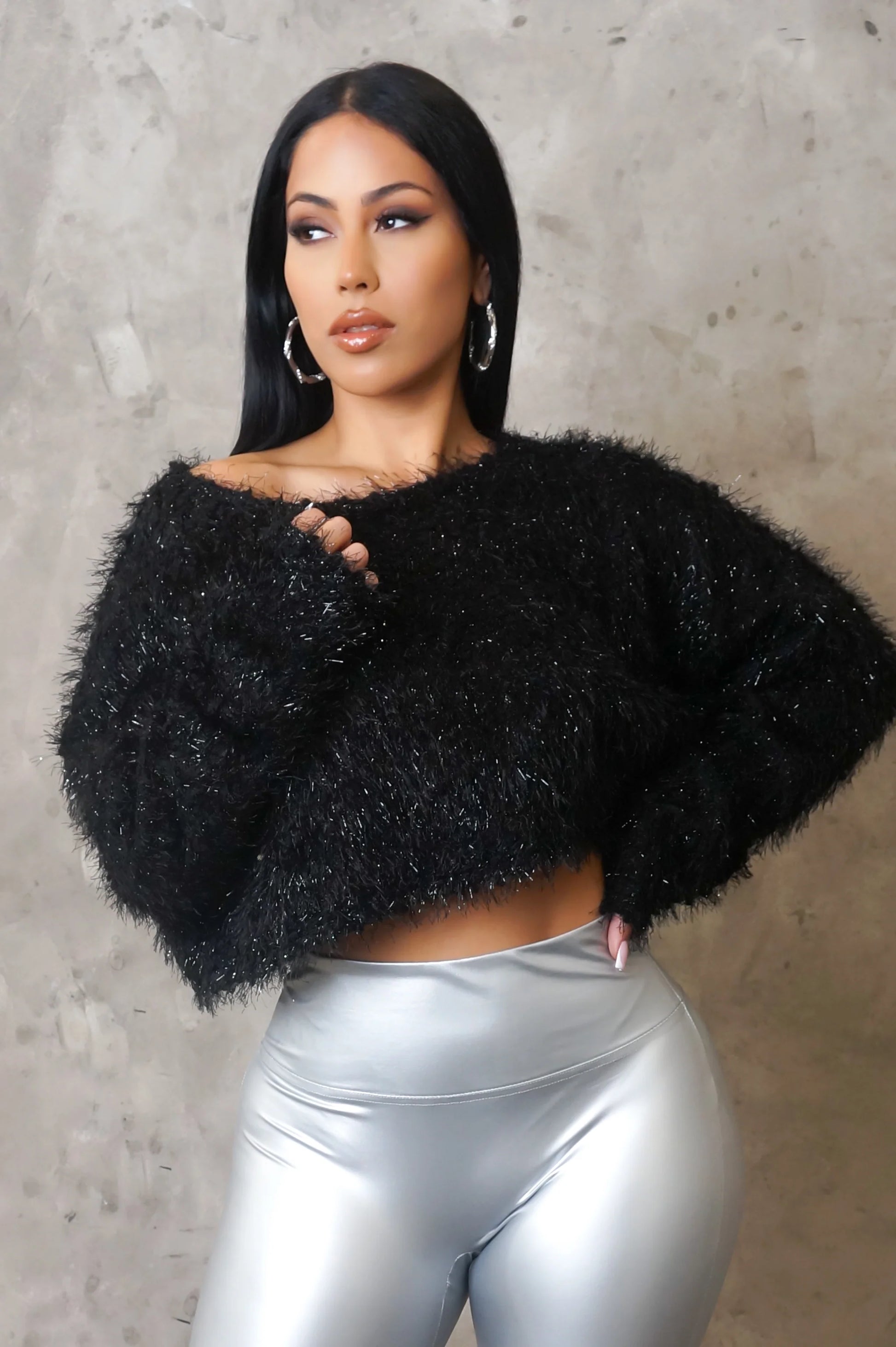 Woman wearing a black fuzzy sweater and silver pants against a gray background