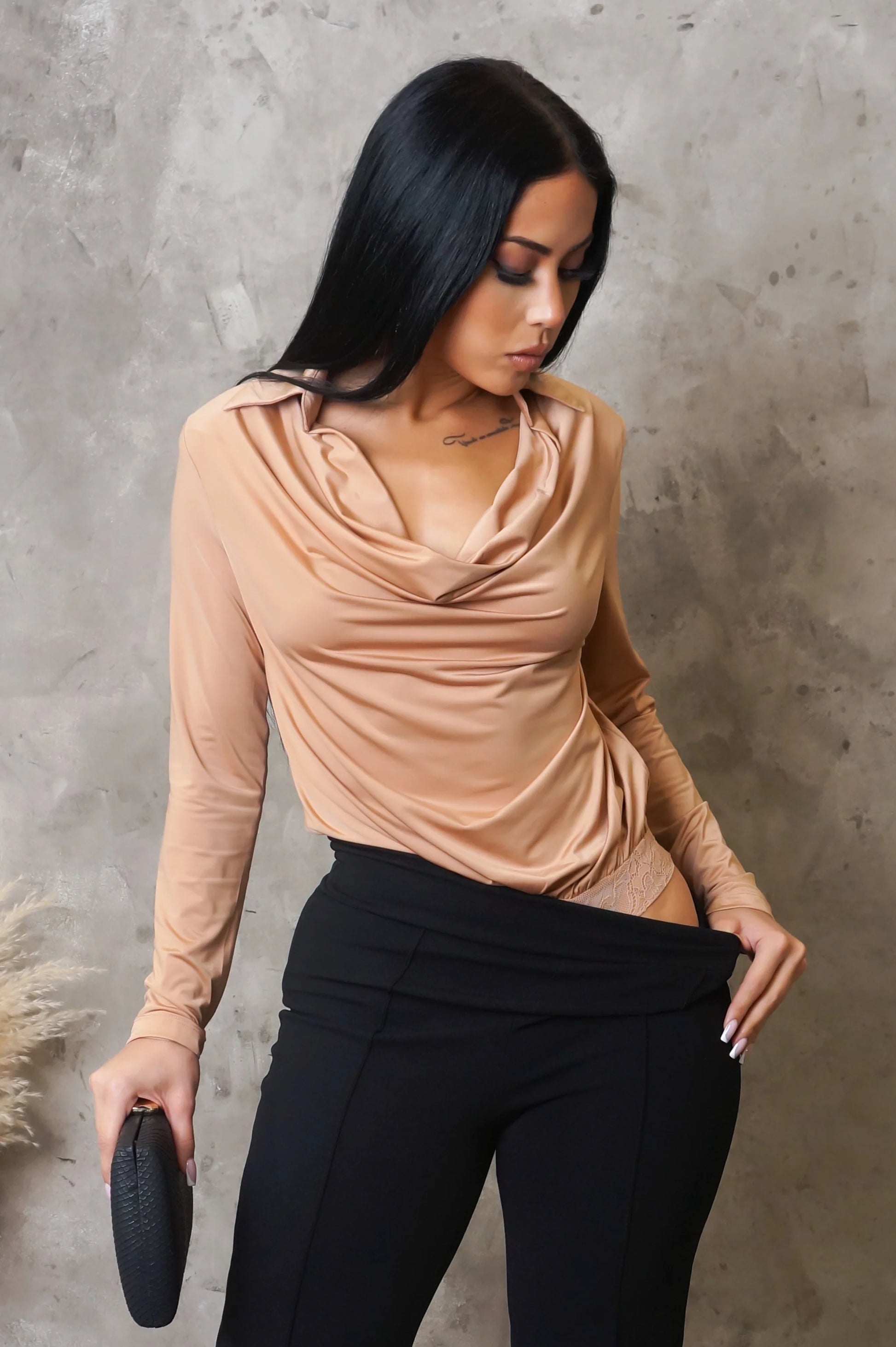 Woman wearing a beige long-sleeve top and black pants against a gray concrete wall.