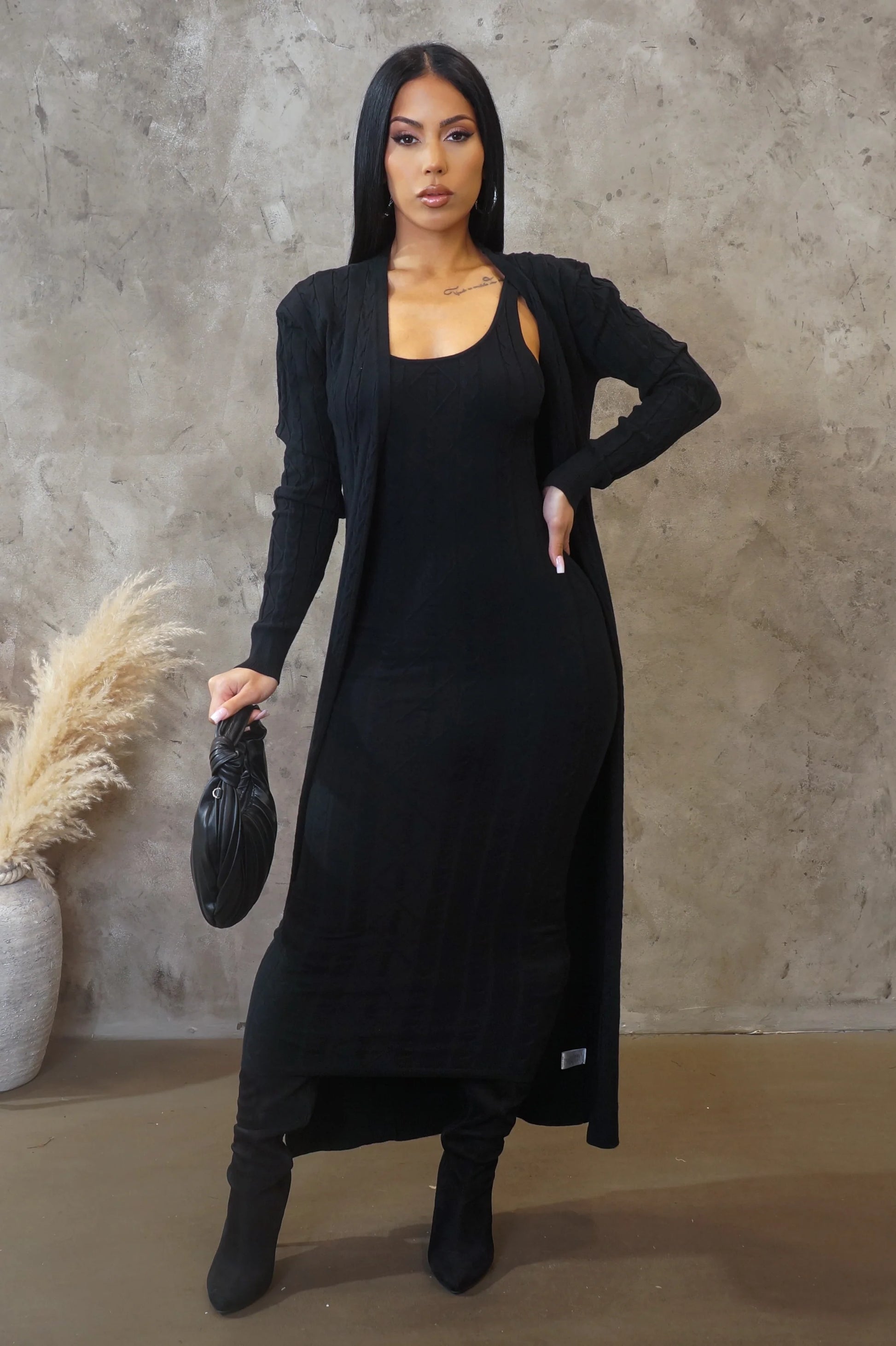 Woman in a black outfit holding a black handbag against a textured wall.