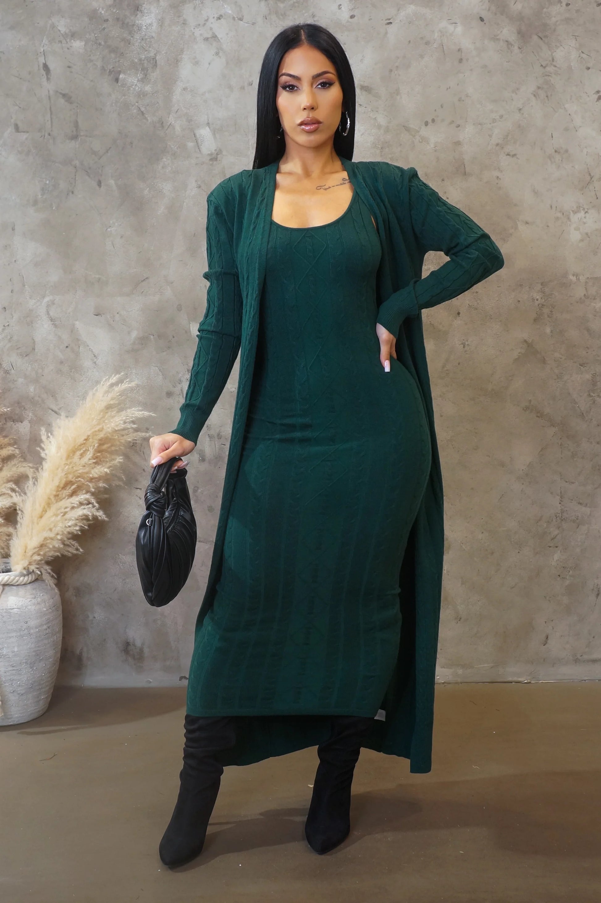 Woman wearing a green dress and cardigan set with a black handbag against a textured wall.