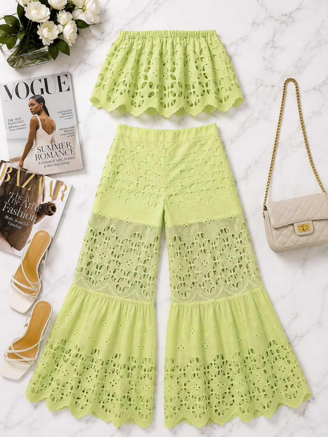 A two-piece women's outfit consisting of a ruffled eyelet crop top and matching wide-leg pants in a light lime green color, styled in a flat lay on a white marble background with a beige quilted handbag, white heels, and Vogue magazines.