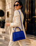 A woman in a cream-colored blazer and trousers walks while wearing a bright blue crocodile-embossed handbag with a top handle and shoulder strap, set against a luxury building entrance.
