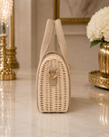 A small woven straw handbag in a warm beige color, standing upright on a polished marble surface. The bag features a structured wicker body, a single top handle, a gold-tone D-ring closure, and a small beige leather accent tab.