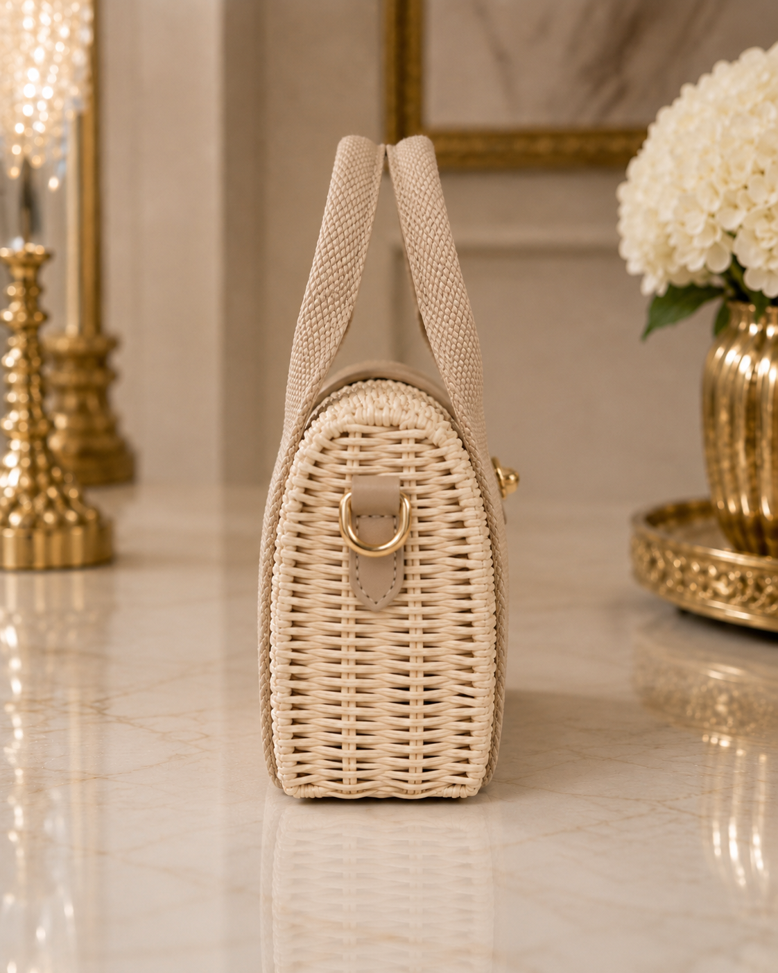 A small woven straw handbag in a warm beige color, standing upright on a polished marble surface. The bag features a structured wicker body, a single top handle, a gold-tone D-ring closure, and a small beige leather accent tab.
