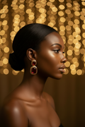 A woman in profile wearing large rectangular gold-toned earrings with deep red gemstones, set against a warm golden bokeh background.