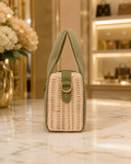 A small woven wicker handbag with an olive green fabric handle and trim, standing on a polished marble surface in a warm, upscale interior setting.