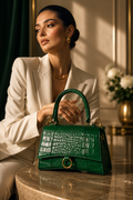 A woman in a cream blazer sits beside a marble table holding a structured emerald green crocodile-embossed handbag with gold-tone hardware.