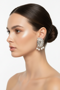 A close-up side profile of a woman with dark hair in a bun, wearing a large, ornate silver-toned statement earring adorned with clear stones and a dangling pearl cluster, against a clean white background.