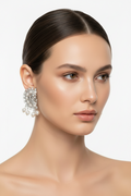 A close-up portrait of a woman with dark hair pulled back, wearing a large ornate silver-toned statement earring with dangling pearl-like accents against a clean light gray background.