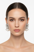 A close-up portrait of a model wearing large, ornate silver-toned statement earrings with clear crystal-like details against a plain light gray background.