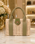 A structured woven straw tote bag with olive green fabric handles and side panels, featuring a central green flap with a gold-tone clasp, displayed on a marble countertop in a boutique setting.