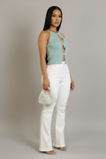A woman stands in a studio wearing a light mint green halter crop top with front cutouts, white high-waisted flared pants, and holding a small white woven handbag against a plain light gray background.