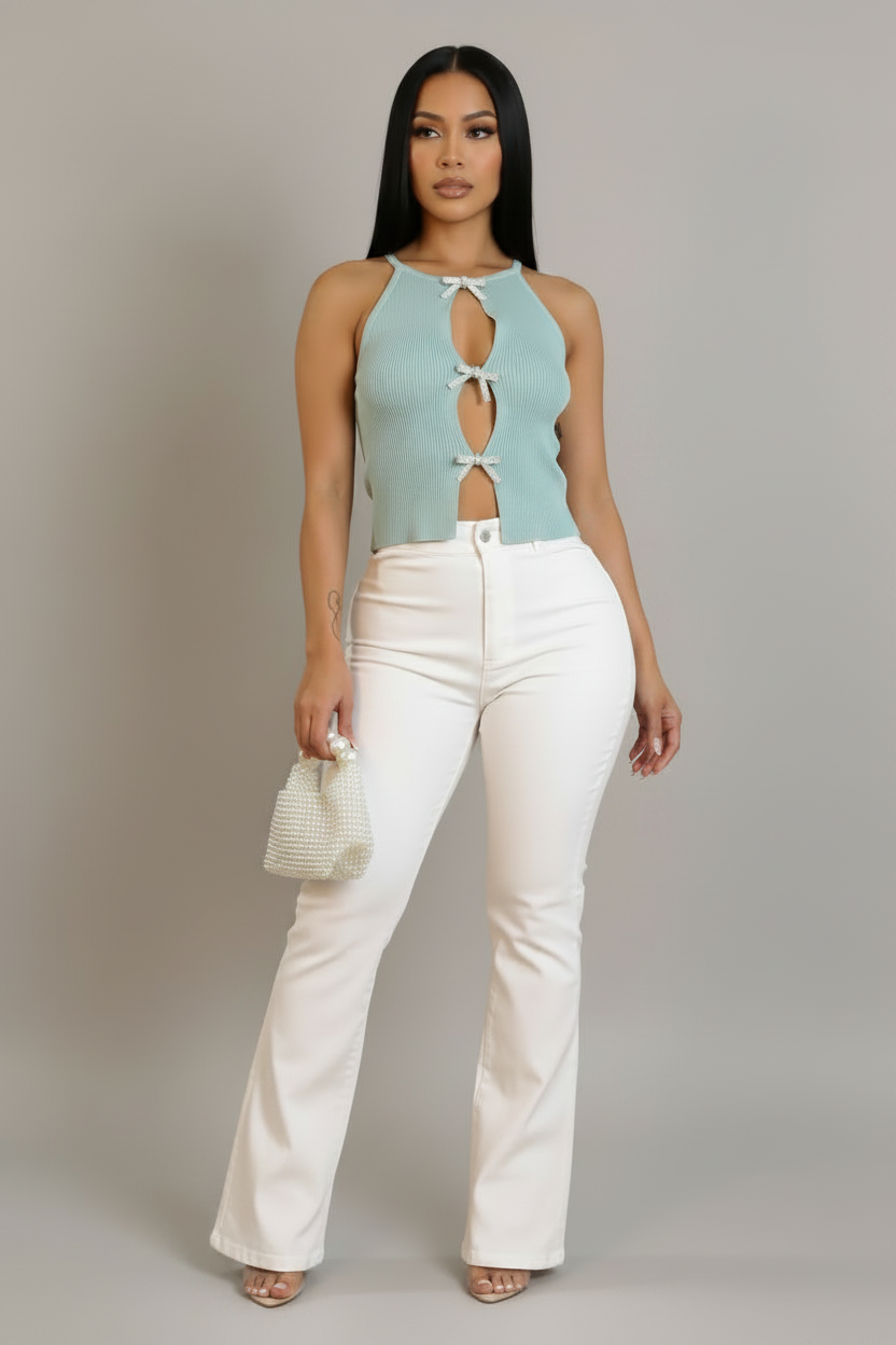 A woman modeling a light blue sleeveless knit top with keyhole cutouts and white high-waisted flare pants, holding a small white woven handbag against a plain light gray studio background.