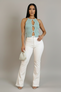 A woman modeling a sleeveless mint green knit halter top with three front keyhole cutouts, paired with white flared pants and holding a small white textured handbag against a plain light gray studio background.