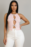 A woman modeling a light pink ribbed halter crop top with keyhole cutouts and bow details, paired with white pants, against a solid gray studio background.