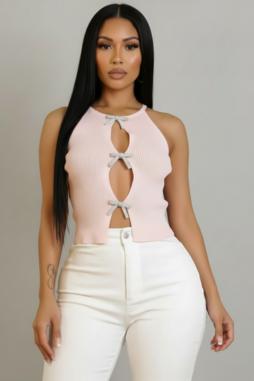 A woman modeling a light pink ribbed halter crop top with three keyhole cutouts and matching bow accents, paired with white pants, against a solid gray studio background.